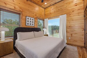 A bedroom with a large bed, wooden walls, a window with blinds, and a framed picture on the wall. - *Gorgeous 5BD Lodge w/ POOL! Hot Tub + Theater* (Sevierville)