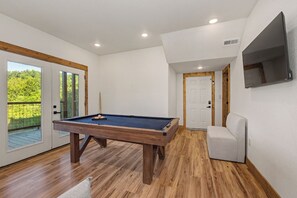 A pool table in a room with wooden flooring, a white wall, and a door leading to a balcony with a view of greenery. - *Gorgeous 5BD Lodge w/ POOL! Hot Tub + Theater* (Sevierville)