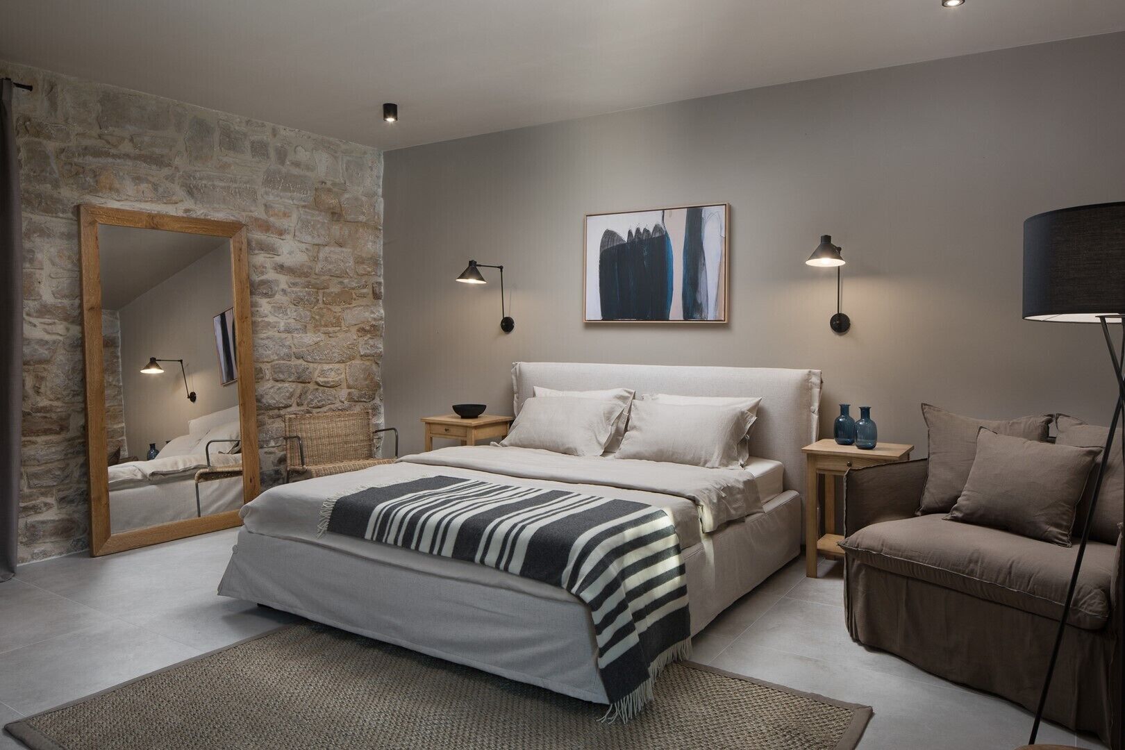 A modern bedroom with a stone wall, a large bed with a striped blanket, two bedside tables with lamps, a chair, and a floor lamp.