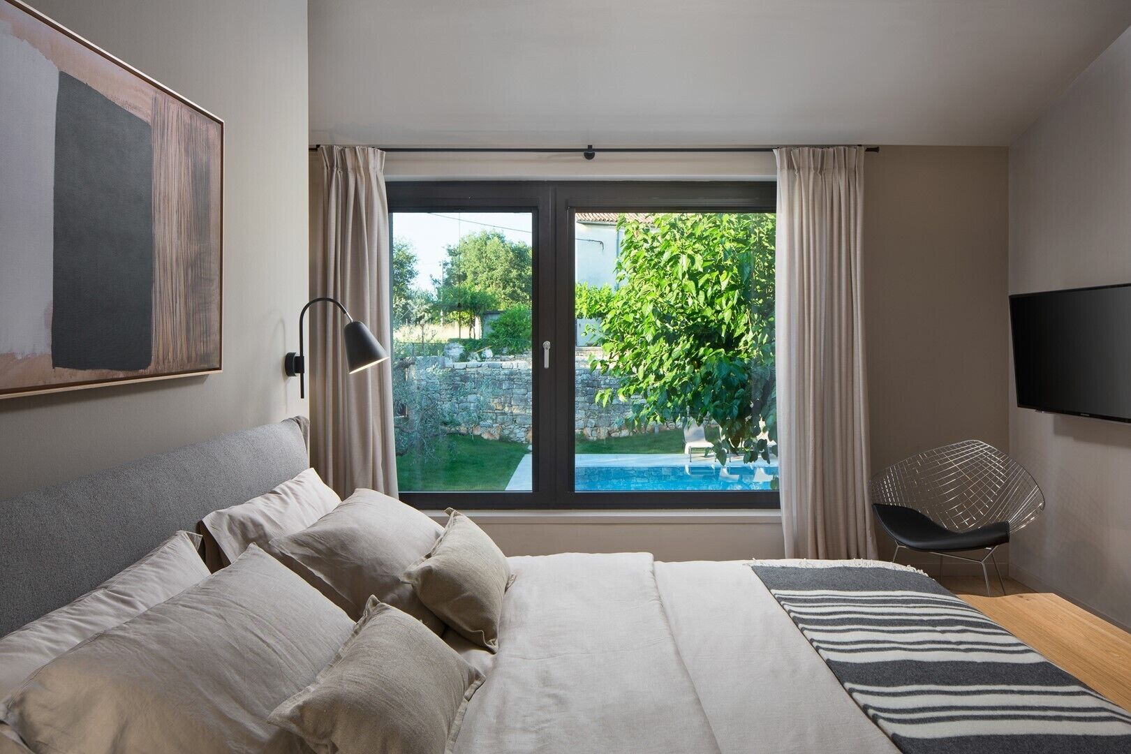 Un dormitorio con cama, televisor, ventana con vista a una piscina y follaje.