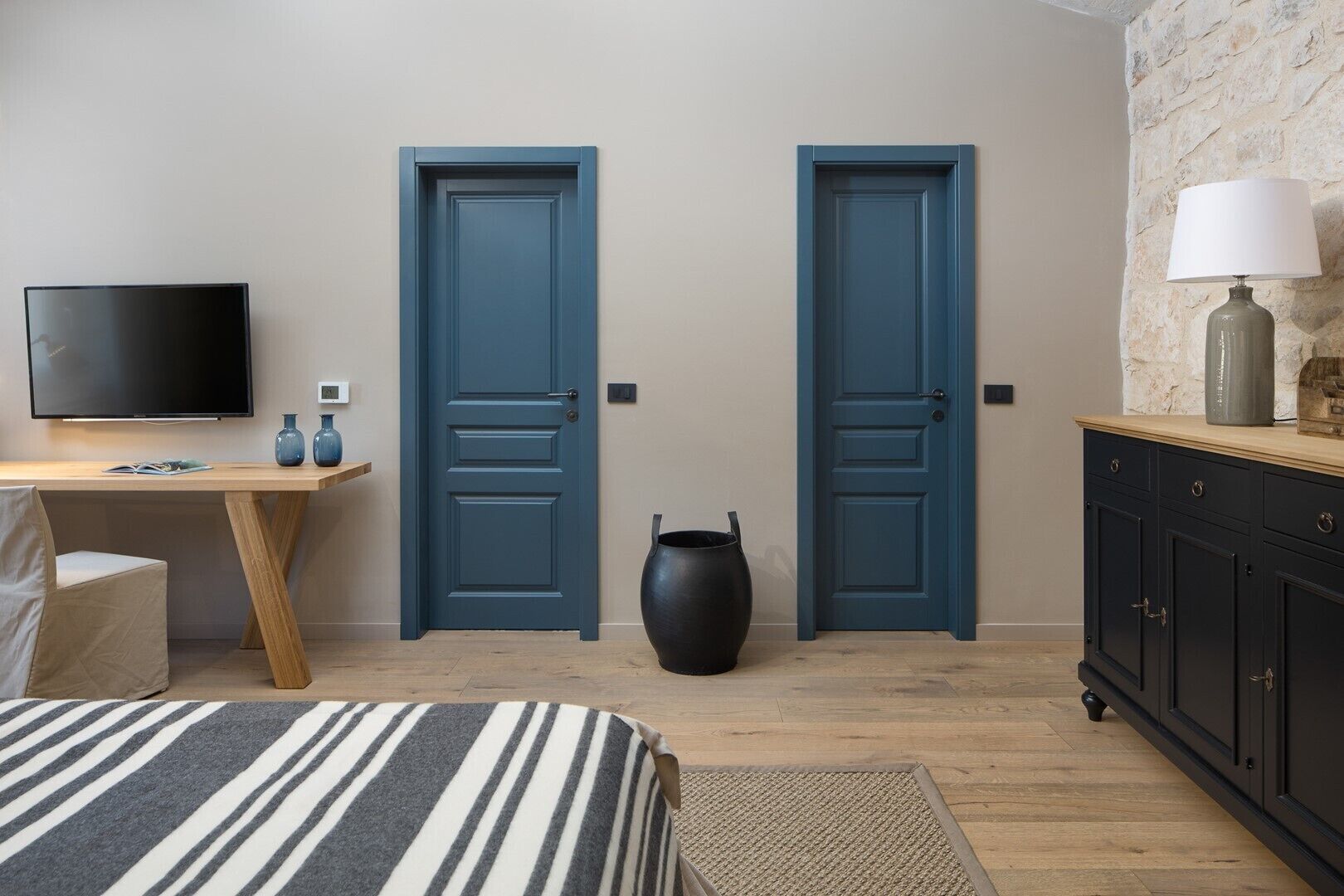 A bedroom with two doors, a TV, a desk, a chair, a bed, a lamp, and a large vase.