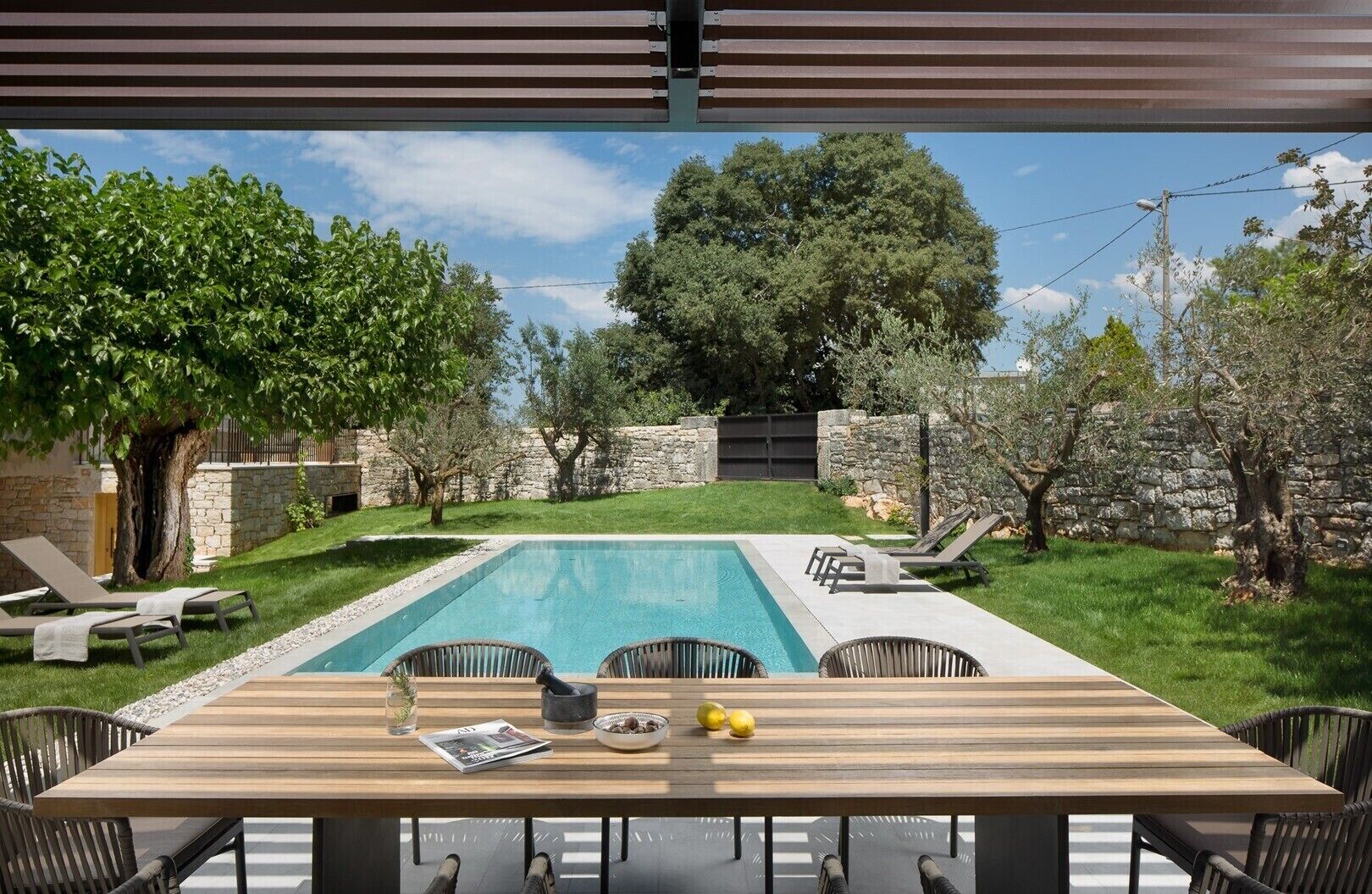 Ein Poolbereich mit Holztisch, Liegestühlen und einer Steinmauer.