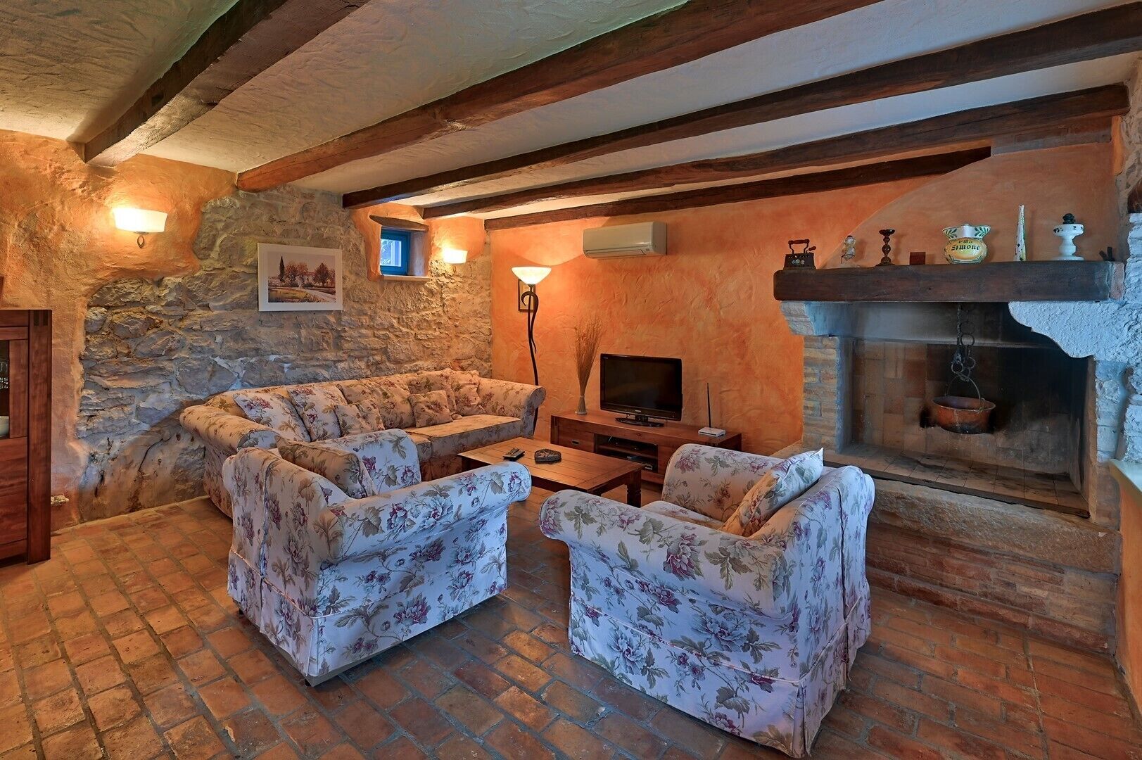 A rustic living room with stone walls, wooden beams, a fireplace, and floral-patterned furniture.
