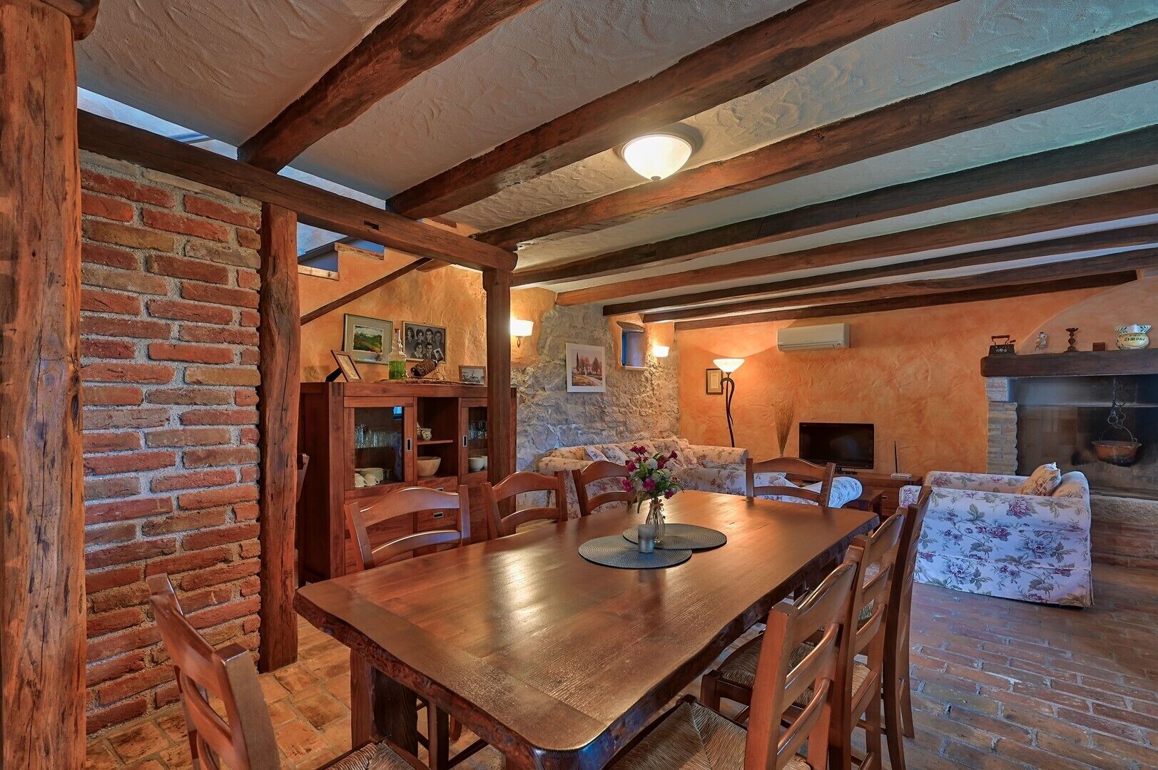 A rustic dining area with a wooden table and chairs, a brick wall, and a stone fireplace.