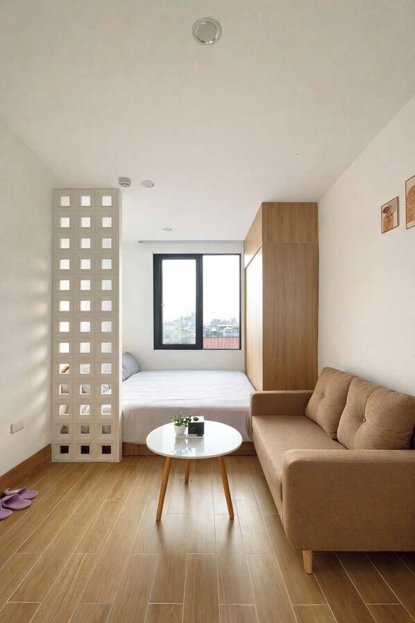 A modern, compact living space with a bed, a sofa, a small table, and a view of the city through a window. - Alley Home & Apartment (Hanoi)