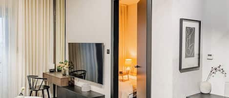 A modern hotel room with a dining area, a large mirror, a bed with a bedside table, and a framed artwork on the wall.
