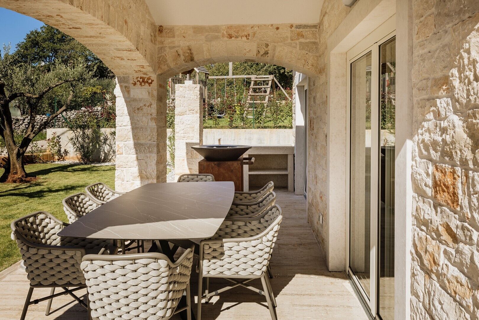 Eine Terrasse im Freien mit einem Steintisch, Korbstühlen, einer Feuerstelle und Blick auf einen Garten und Bäume.
