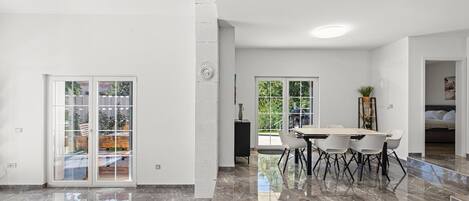Ampla sala de estar com piso de mármore, área de refeições com mesa e cadeiras, e grandes portas de vidro deslizantes que dão para uma varanda.