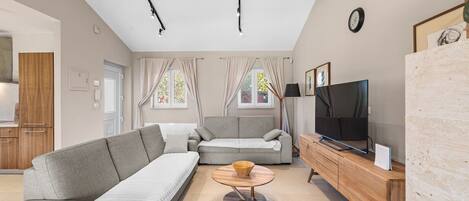 A modern living room with a grey sofa, a wooden coffee table, a TV on a wooden stand, and a kitchen area in the background.