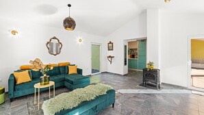 Un salon spacieux avec un canapé de couleur turquoise, une petite table ronde et un pouf vert. Un poêle noir se trouve dans un coin, et une porte verte donne accès à une autre pièce.
