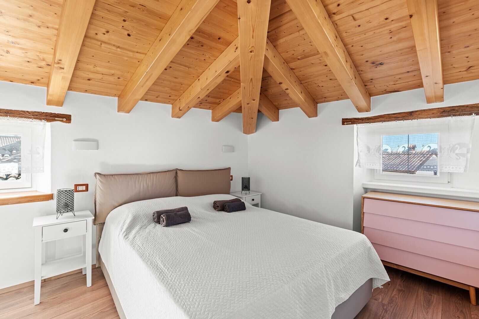 A bedroom with a wooden ceiling, a bed with a white quilt, a bedside table with a lamp, and a dresser with a pink finish.