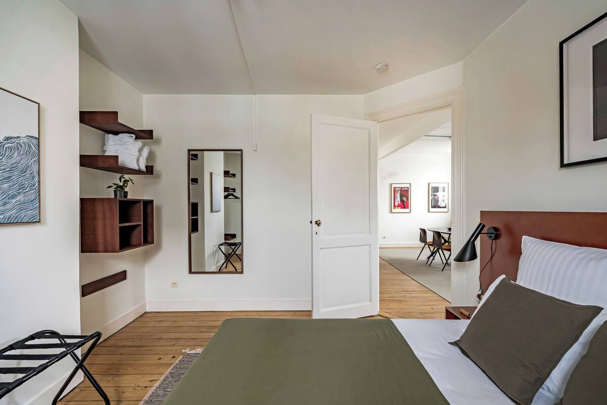 Un dormitorio con cama, espejo, estante con ropa doblada y puerta que da a otra habitación.