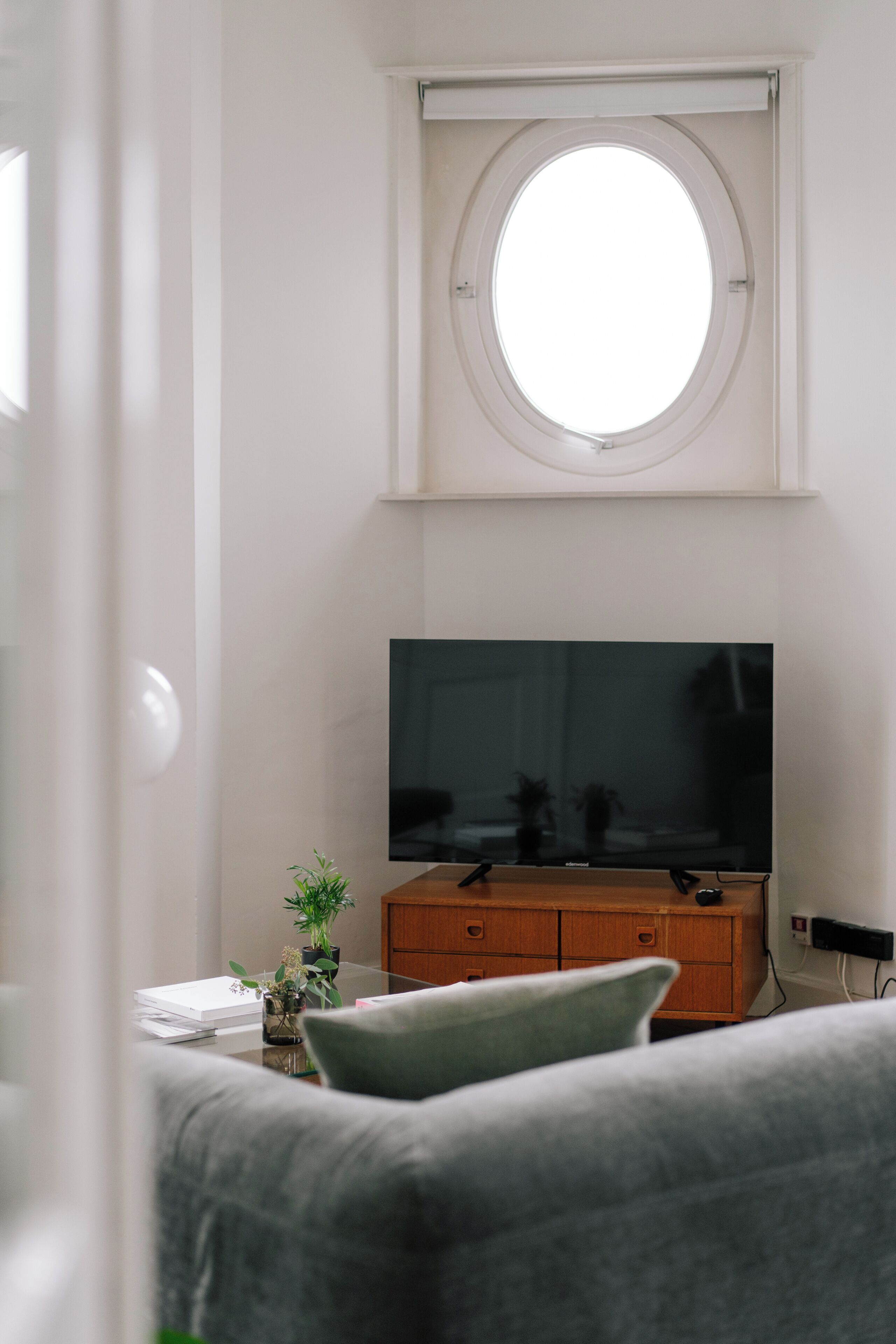 A room with a television on a wooden stand, a round window, and a grey sofa.