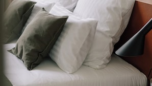 Un lit bien fait, avec des oreillers blancs et gris, une tête de lit en bois et une table de chevet sur laquelle se trouvent une lampe et une bouteille.