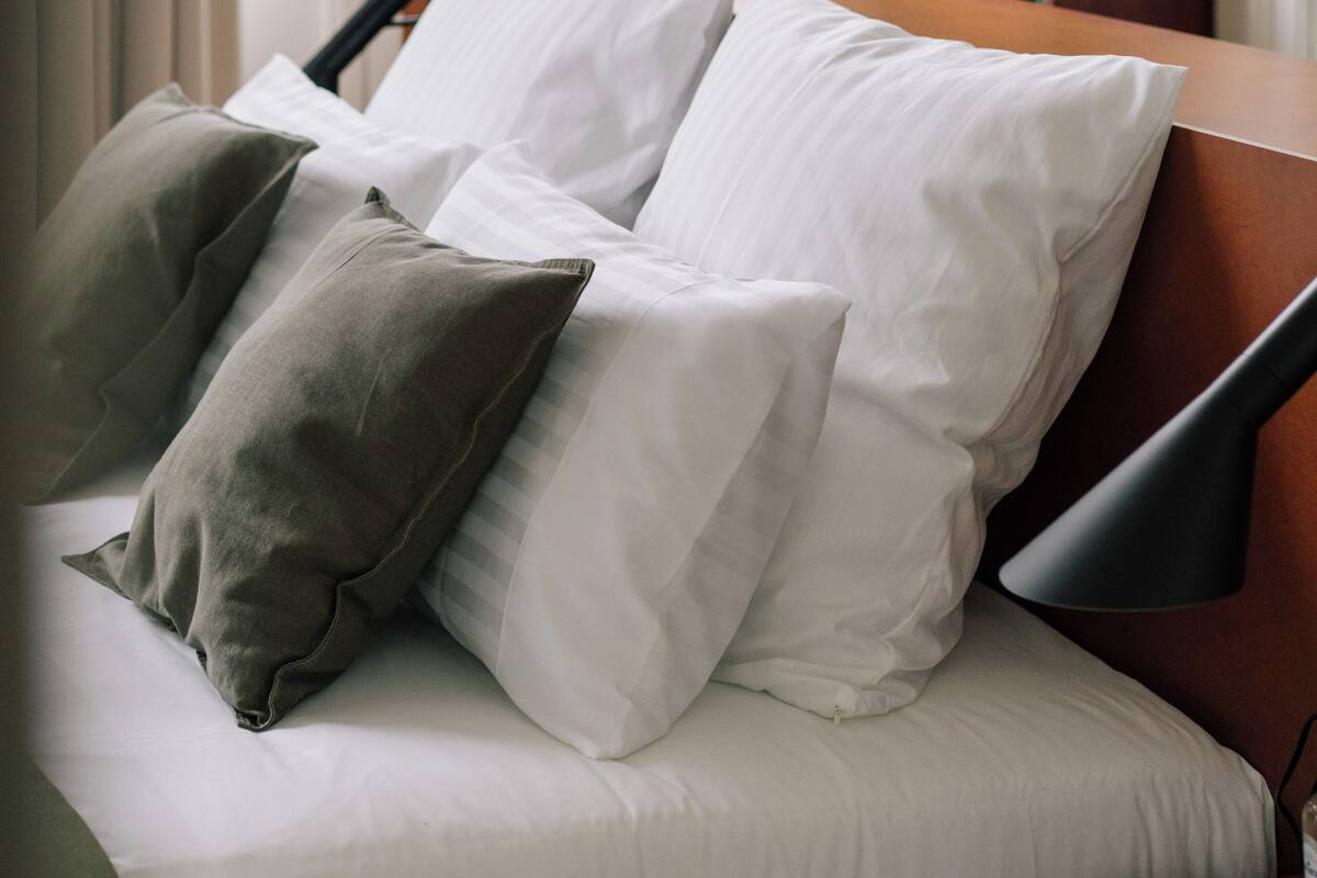 Una cama bien hecha con almohadas blancas y grises, un cabecero de madera y una mesita de noche con una lámpara y una botella.