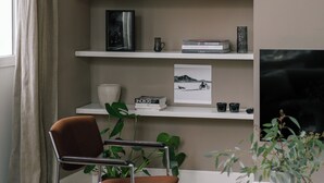 Un salon avec un fauteuil marron, une bibliothèque remplie de divers objets, un téléviseur et une fenêtre avec des rideaux.