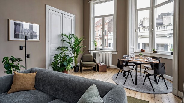 Un salon avec un canapé gris, une table à manger avec des chaises, un radiateur et de grandes fenêtres donnant sur la ville.