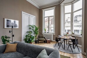 A living room with a grey sofa, a dining table with chairs, a radiator, and large windows with a city view. - A Place - Schuttershofstraat 1 (Antwerp)