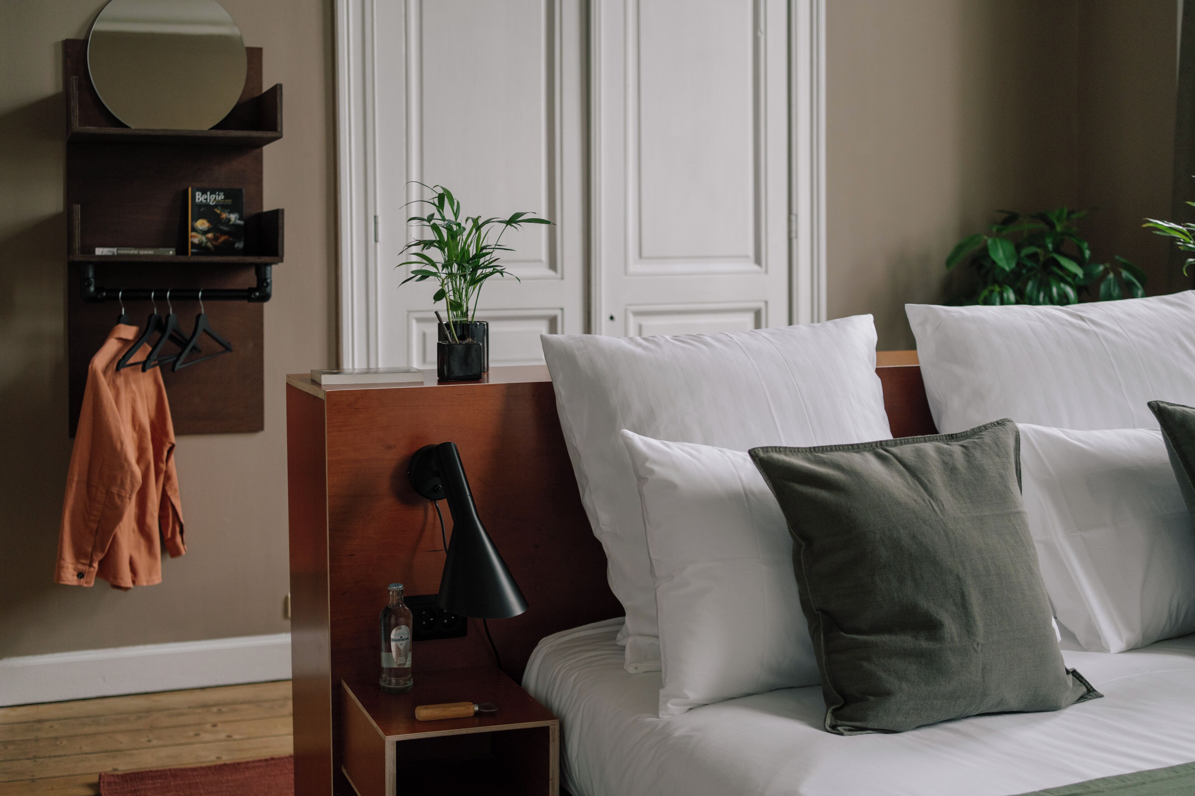 A neatly arranged hotel room with a bed, bedside table, lamp, and a shelf with a book and a plant.
