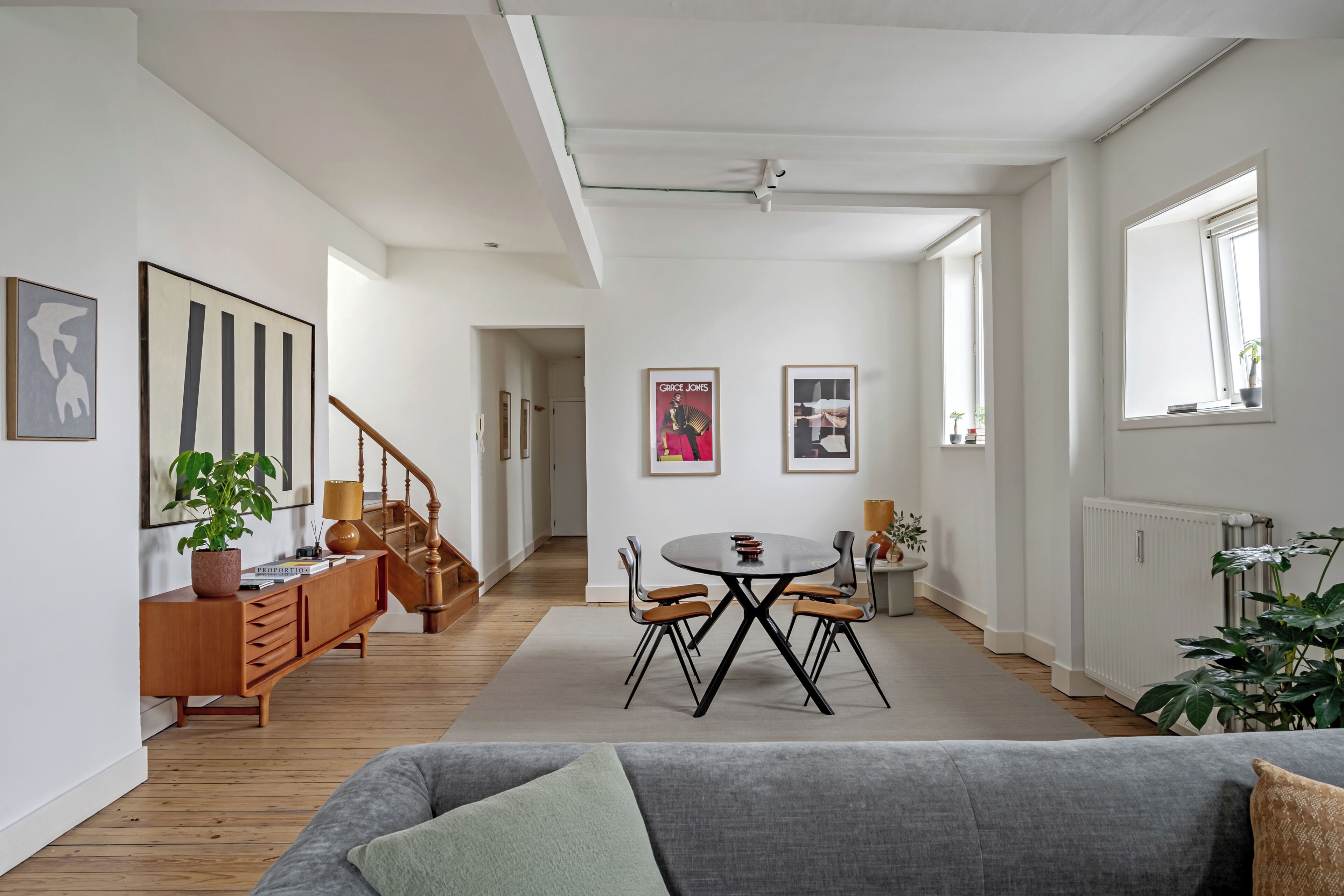 Ein modernes Wohnzimmer mit grauem Sofa, Holzboden, einem Esstisch mit Stühlen, einer Treppe und einer Heizung.