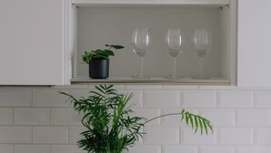 Une cuisine avec un plan de travail blanc, un tableau encadré, une plante en pot et trois verres à vin posés sur une étagère.