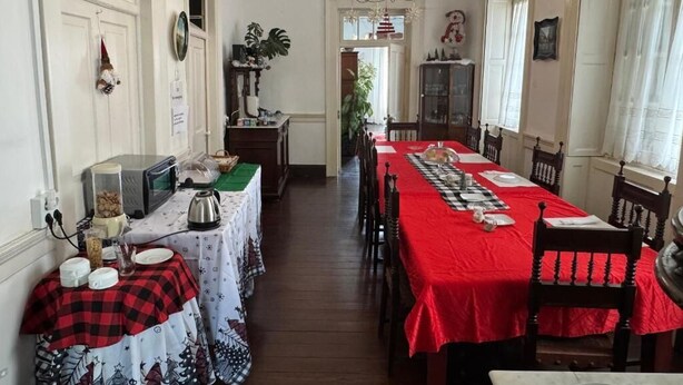 A dining area with a long table covered in a red tablecloth, flanked by wooden chairs. A chandelier hangs from the ceiling, and there are various kitchen appliances and items on the side tables. - VCGuesthouse Grandfather's Room (Funchal)