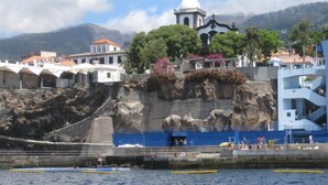 A coastal town with a prominent church, rocky cliffs, and a pier with people and boats. - VCGuesthouse Grandfather's Room (Funchal)