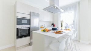 Eine moderne Küche mit weißen Schränken, einem Einbauofen, einem Kühlschrank und einem Essbereich mit weißen Stühlen sowie einer kleinen Kochinsel mit Früchten und einer Kaffeemaschine.