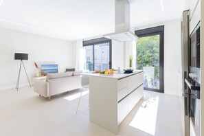 A modern kitchen with a central island, a dining area with a sofa, and a large window providing a view of greenery. - Suites Brisamar III by VillaGranCanaria (Las Palmas de Gran Canaria)