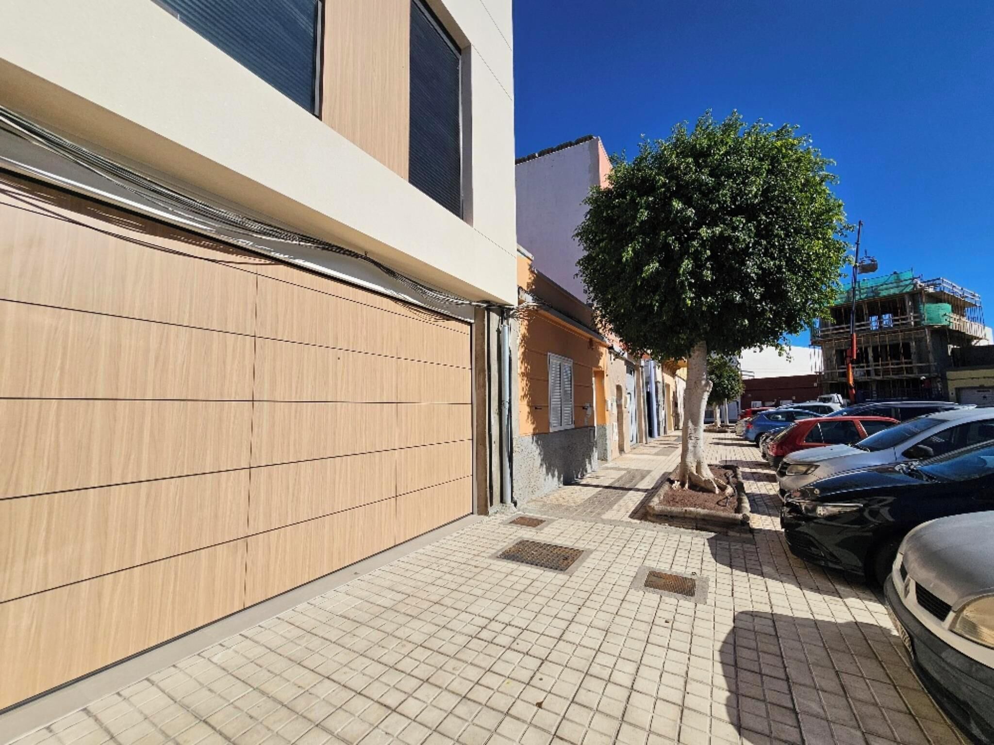 Una calle estrecha con coches estacionados, aceras arboladas y edificios con contraventanas cerradas.
