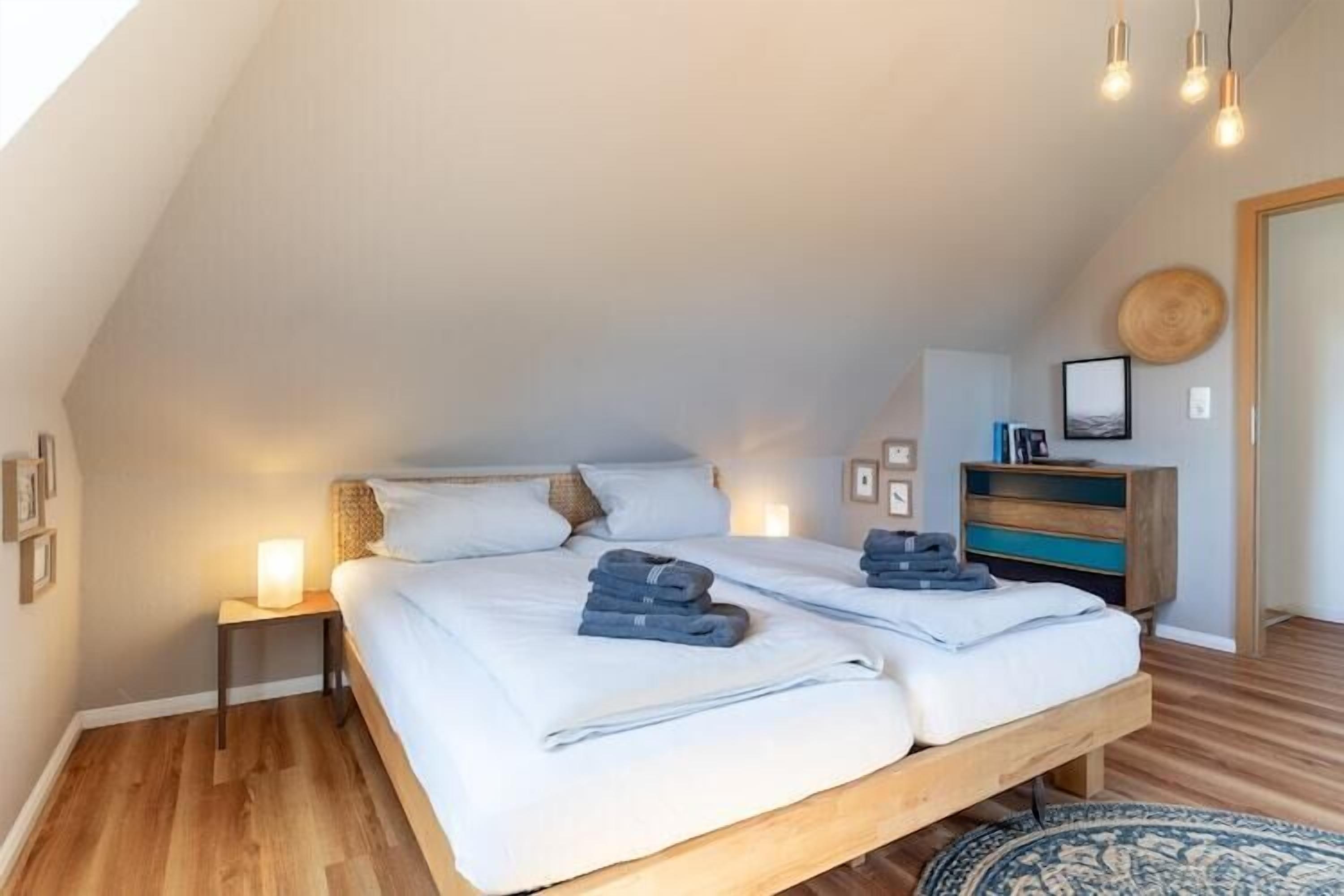 Quarto com cama de madeira, mesas de cabeceira com luminárias, cômoda e tapete redondo.
