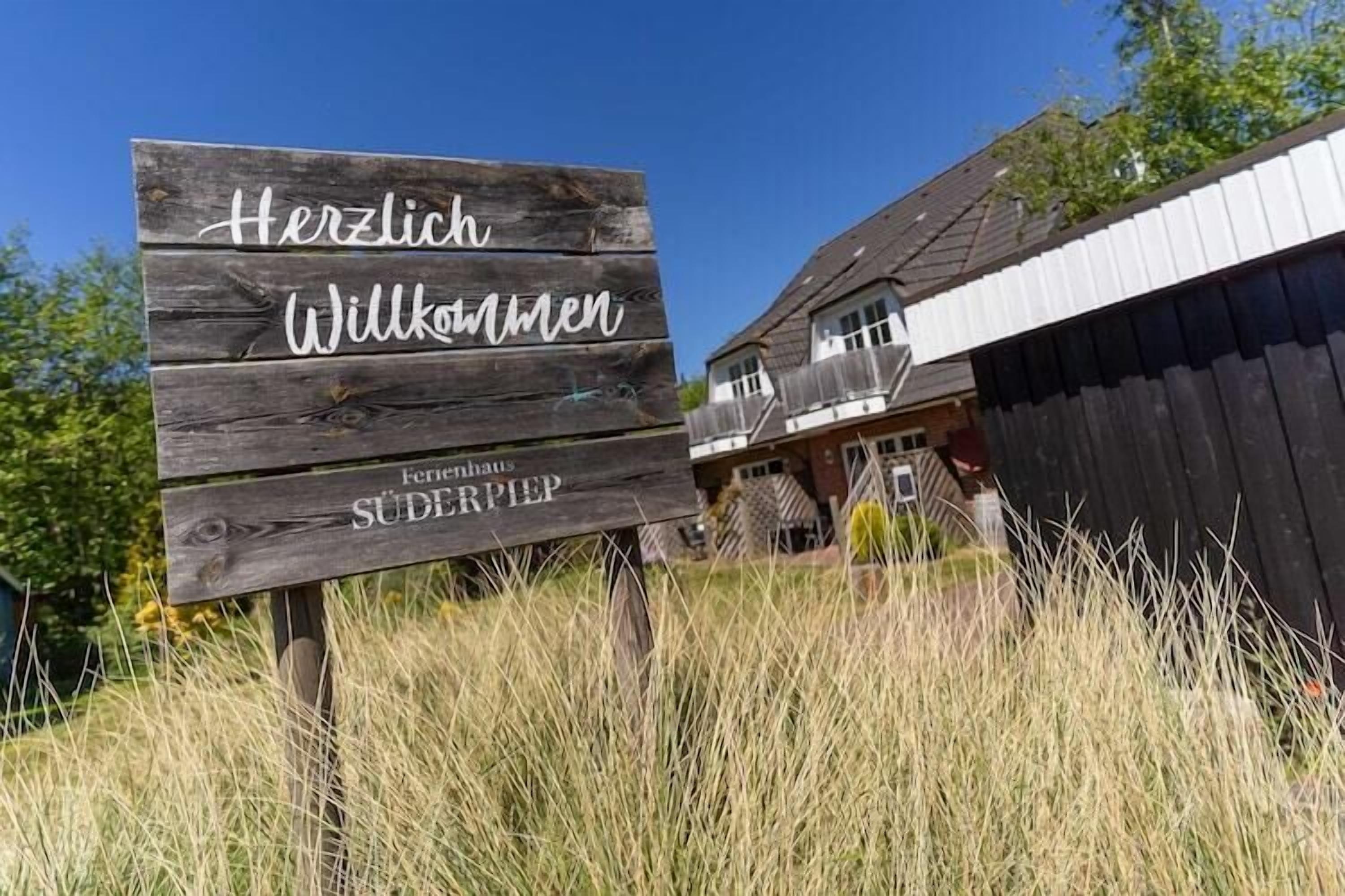 A wooden sign with "Herzlich Willkommen" and "Fernhaus Süderpfiep" written on it, placed in front of a building with a sloped roof.