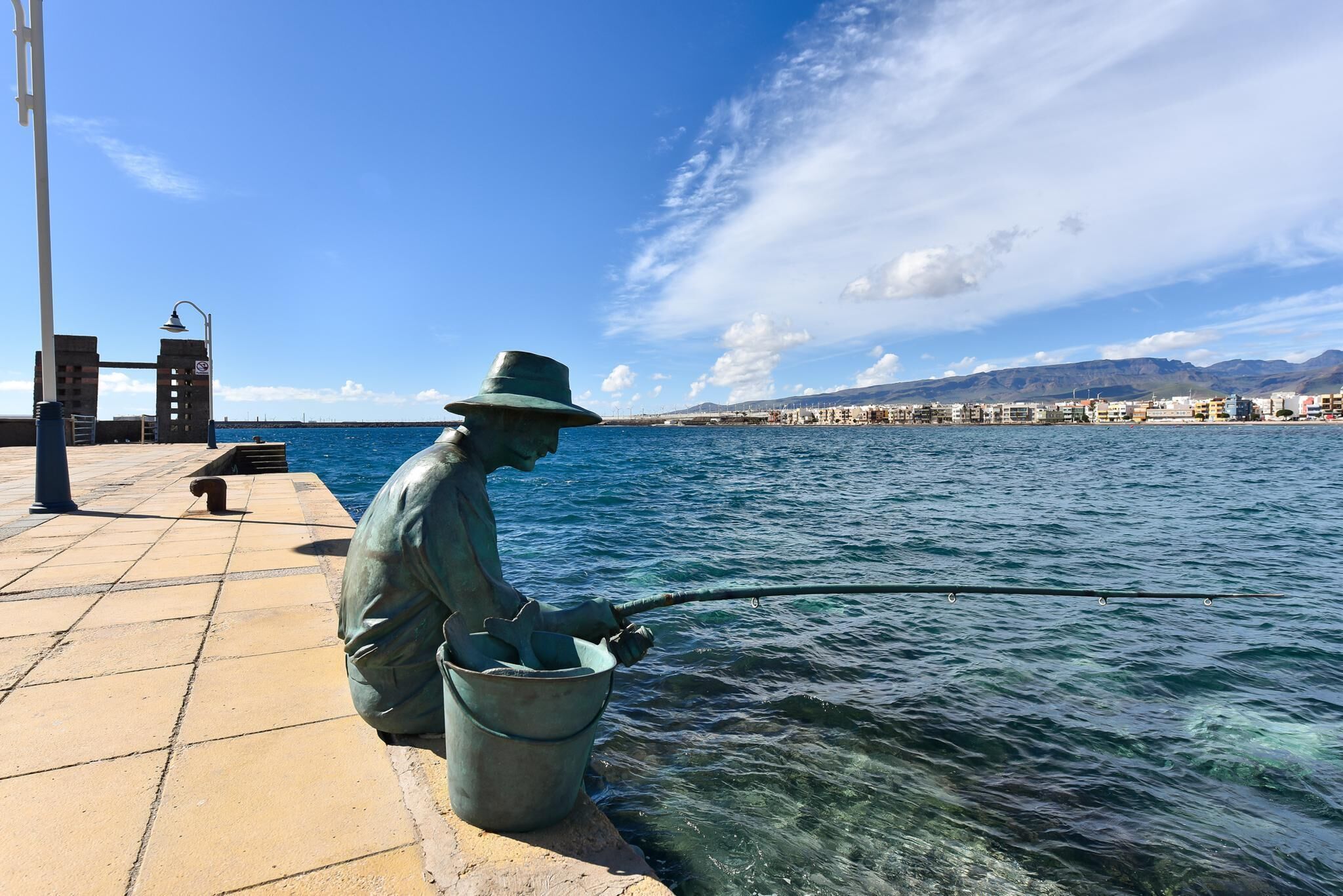 A statue of a fisherman sitting on a bench by the water.