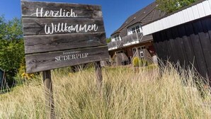 Een houten bord met de tekst 'Herzlich Willkommen' en 'Fernhaus Süderpfiep', geplaatst voor een gebouw met een schuin dak.