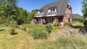 Een twee verdiepingen tellend huis met een schuin dak, een veranda en een kleine tuin met verschillende planten en een pad naar de ingang.