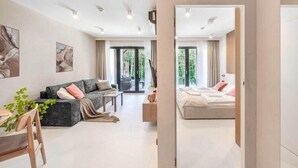 Een moderne woonkamer met een bank, salontafel en een eethoek. Er is een bed met hoofdeinde en een raam met gordijnen.