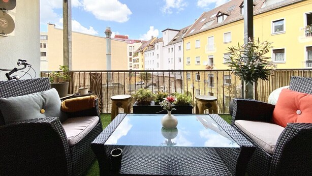 Ein Balkon mit Korbmöbeln, einem Glastisch und Topfpflanzen.