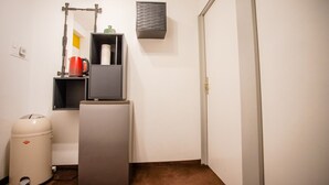 Ein kleines Zimmer mit Kühlschrank, Mülleimer, Ablagefläche mit Wasserkocher und Wandregal.