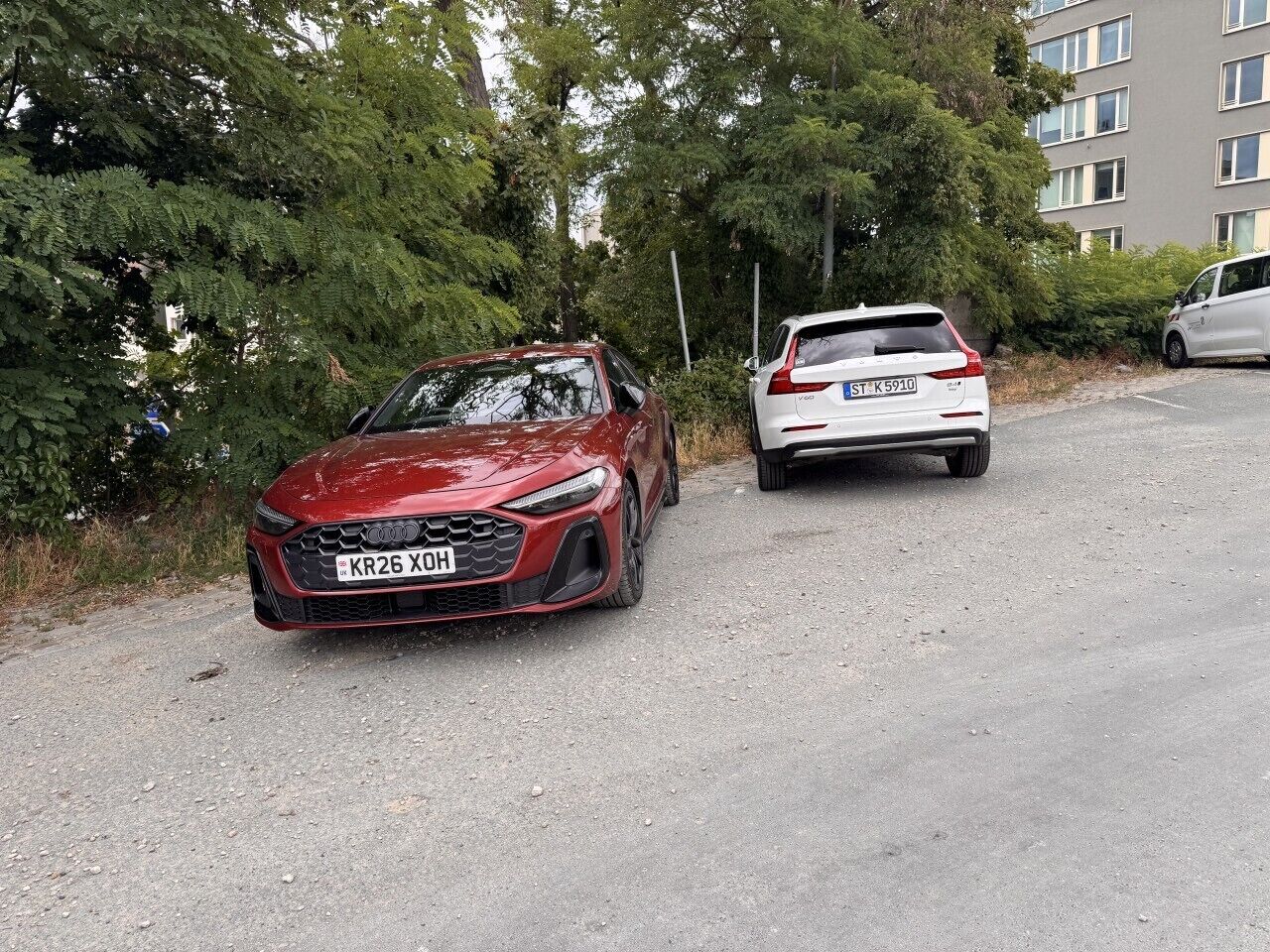 Ein rotes Auto steht auf einem Schotterweg, dahinter ein weißes Auto.