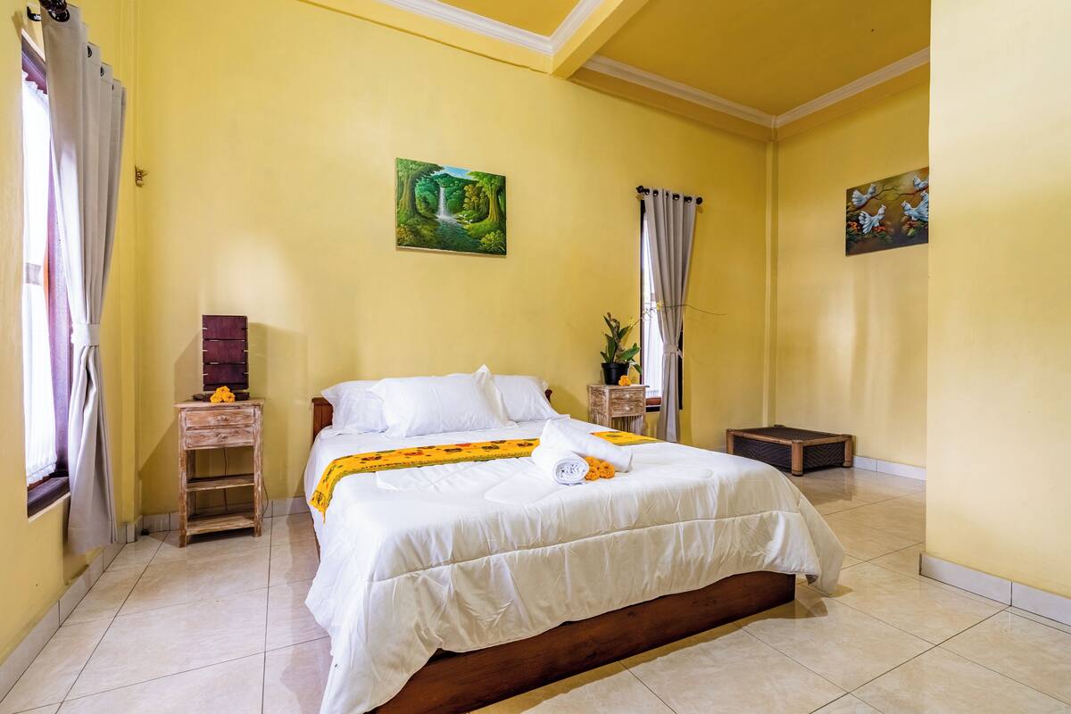 Een slaapkamer met een bed, een nachtkastje, een klein houten kastje, een raam met gordijnen en een schilderij aan de muur.