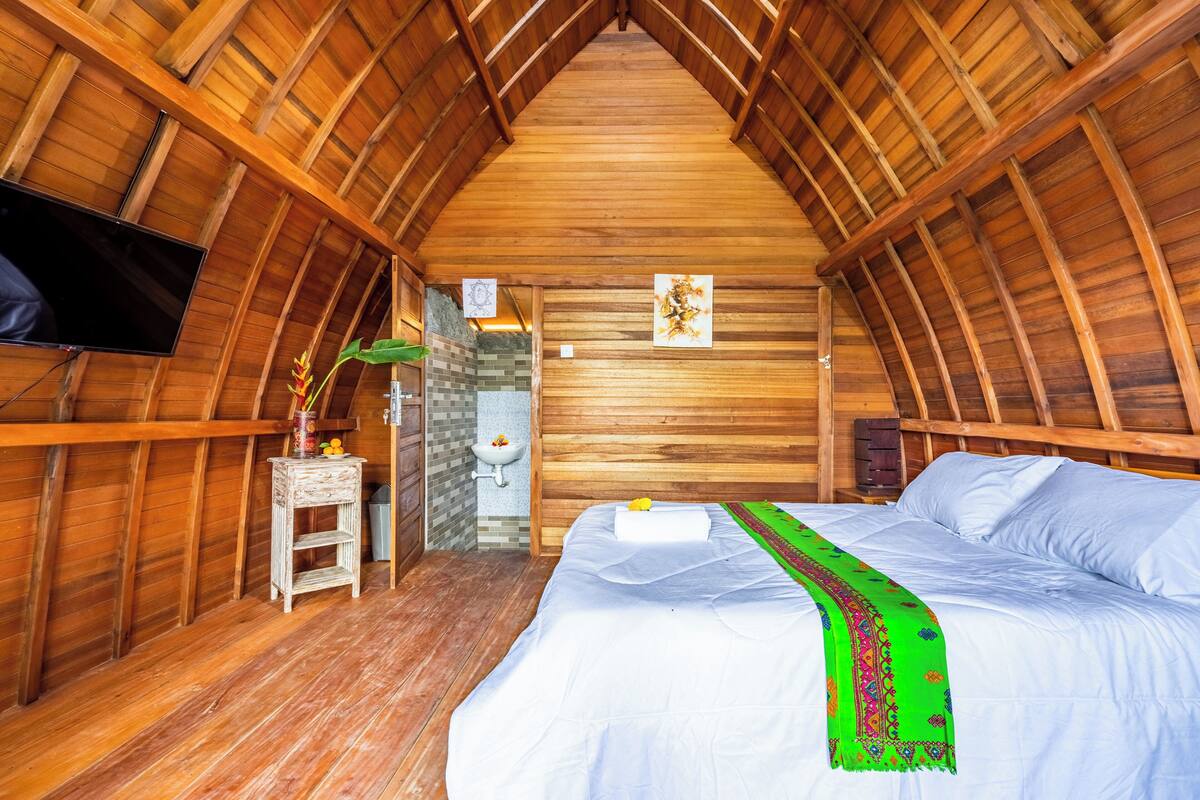 Een knus zolderkamer met houten plafond, een bed met wit beddengoed, een groene plaid, een klein nachtkastje met een vaas, en een tv aan de muur.