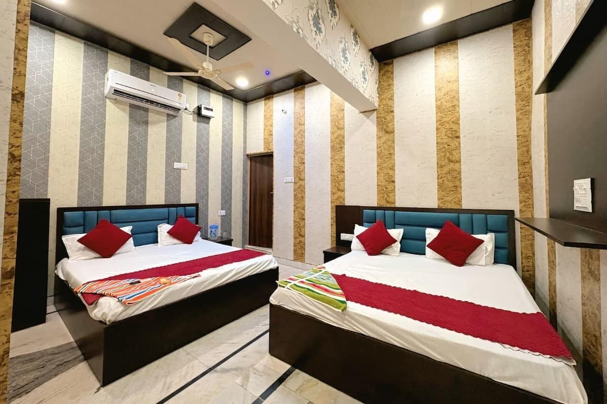 A hotel room with two beds, each with a blue headboard, white bedding, and red pillows. There is a ceiling fan, an air conditioning unit, and a patterned wall.