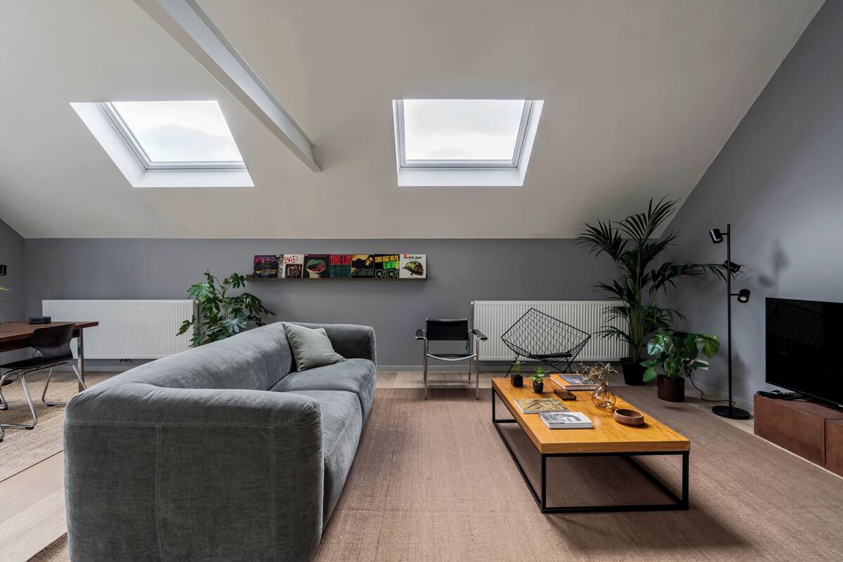 Ein modernes Wohnzimmer im Loft-Stil mit grauem Sofa, einem Holztisch, einem schwarzen Sessel und einem großen Fernseher. Im Deckenbereich befinden sich Dachfenster, ein Heizkörper und ein Blumentopf.