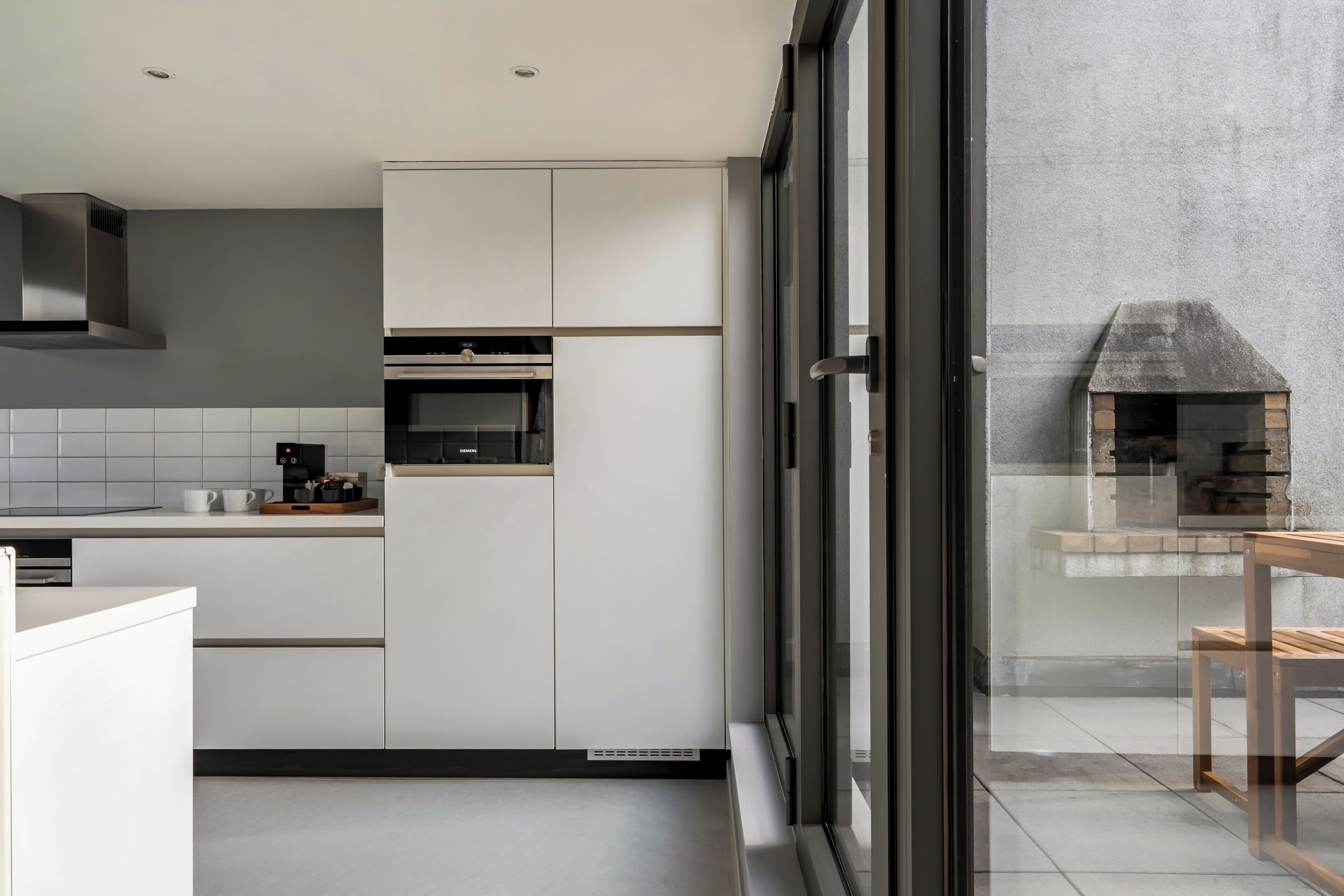 A modern kitchen with white cabinetry, a built-in oven, and a glass-enclosed outdoor cooking area.