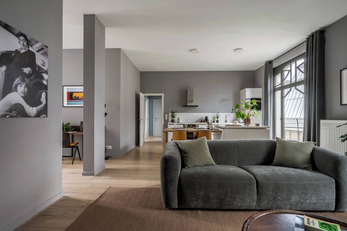 Ein modernes Wohnzimmer mit grauem Sofa, Essbereich, Küche und großen Fenstern.