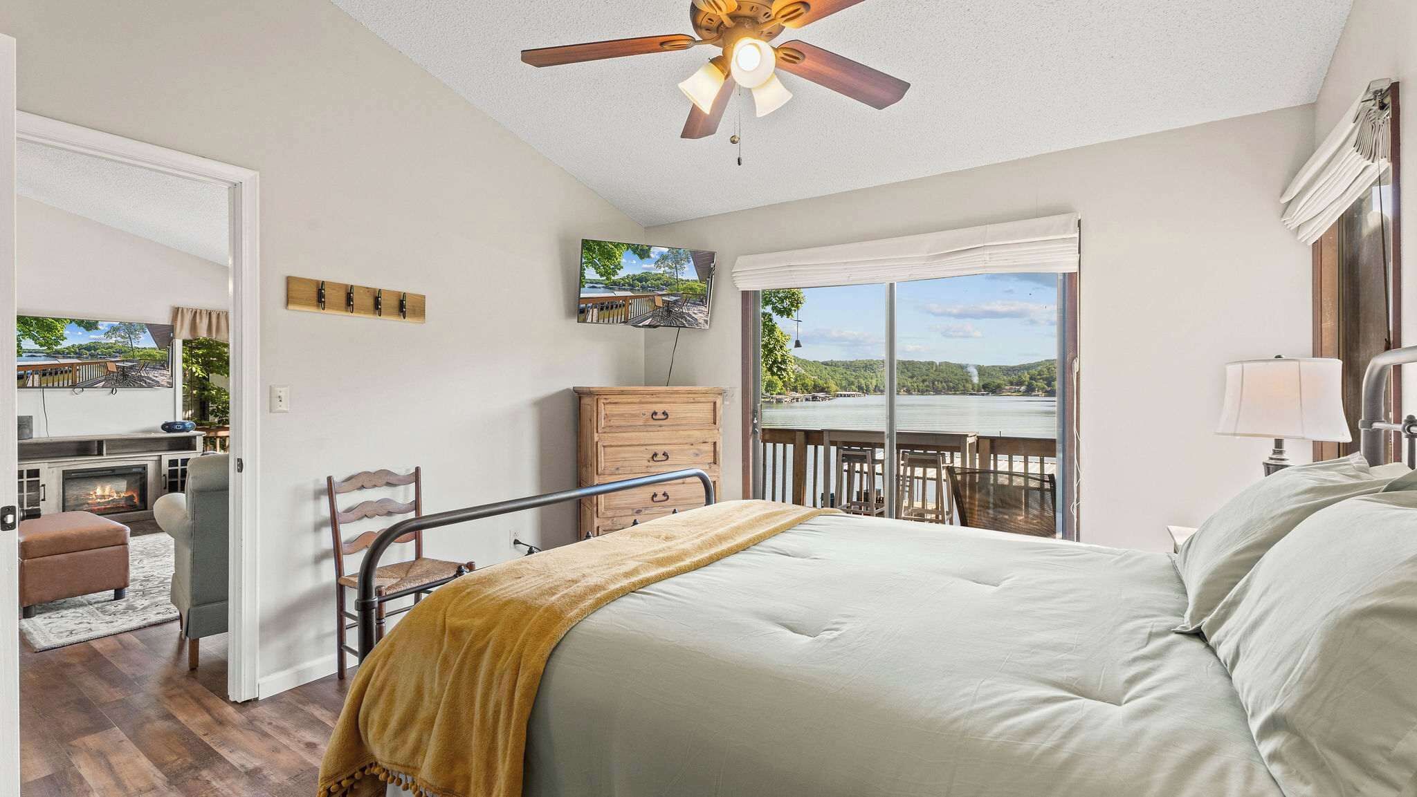 A bedroom with a bed, a ceiling fan, a dresser, a chair, a TV, and a view of a lake through large windows.