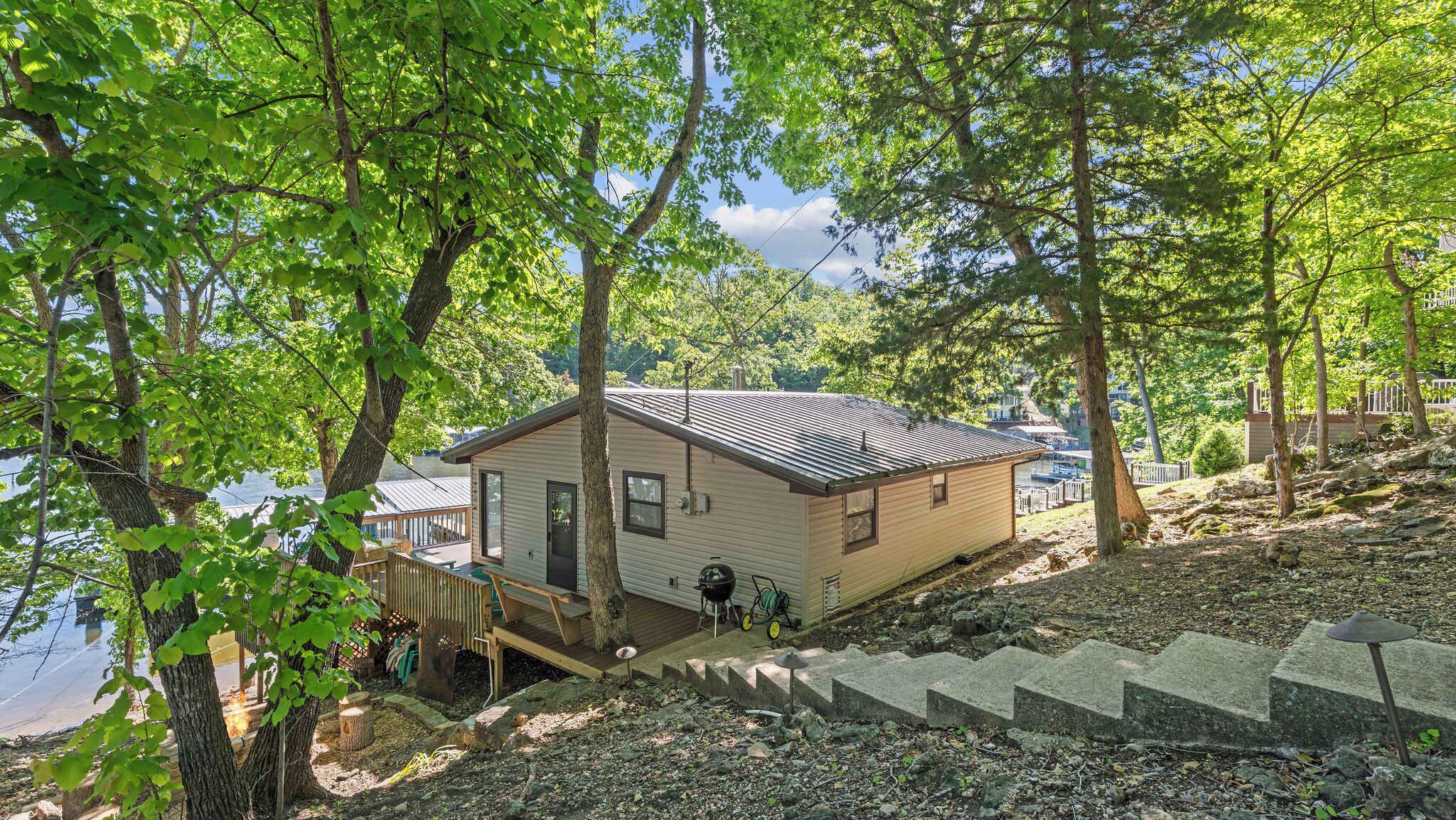 A small cabin with a deck, near a body of water, surrounded by trees.