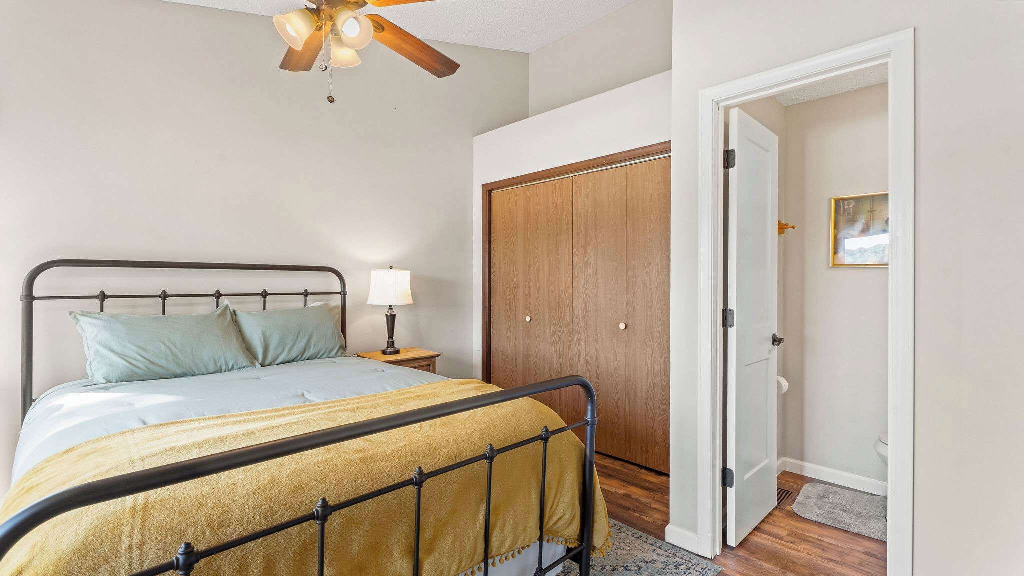 A bedroom with a bed, bedside table, lamp, wooden wardrobe, and a door leading to another room.