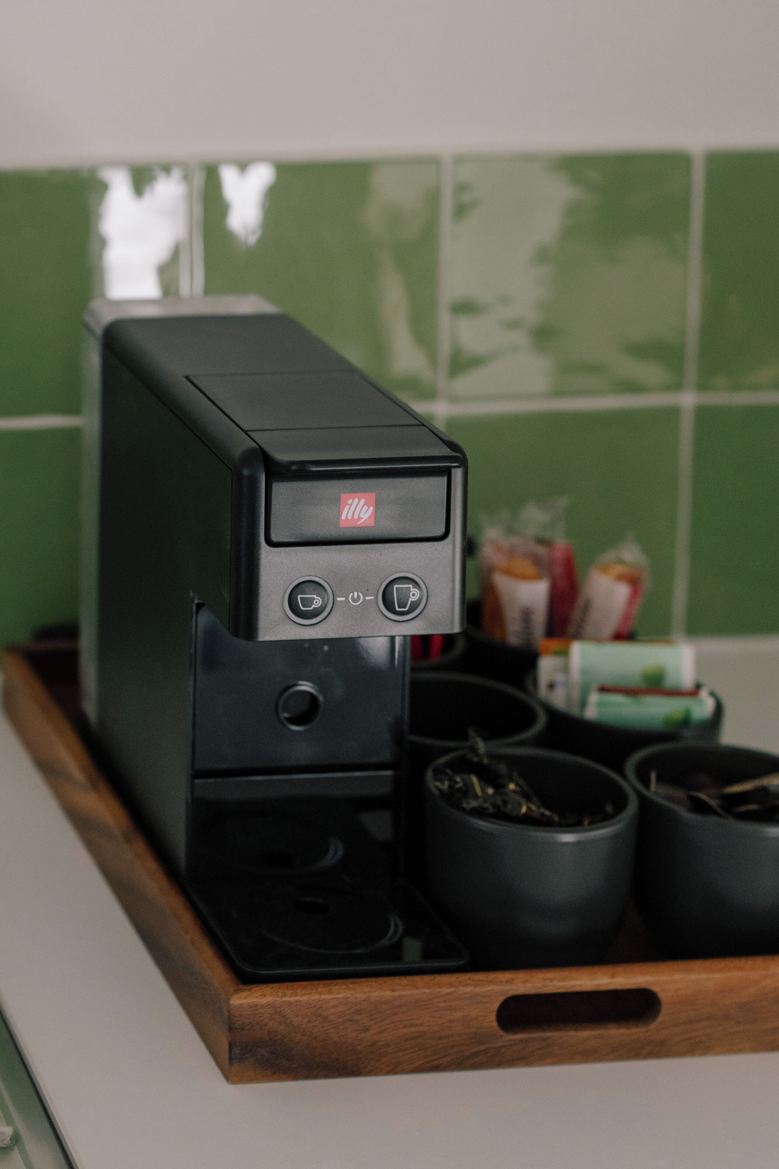 Een koffieapparaat op een houten dienblad, met drie zwarte kopjes, een groene betegelde muur en enkele verpakte etenswaren op de achtergrond.