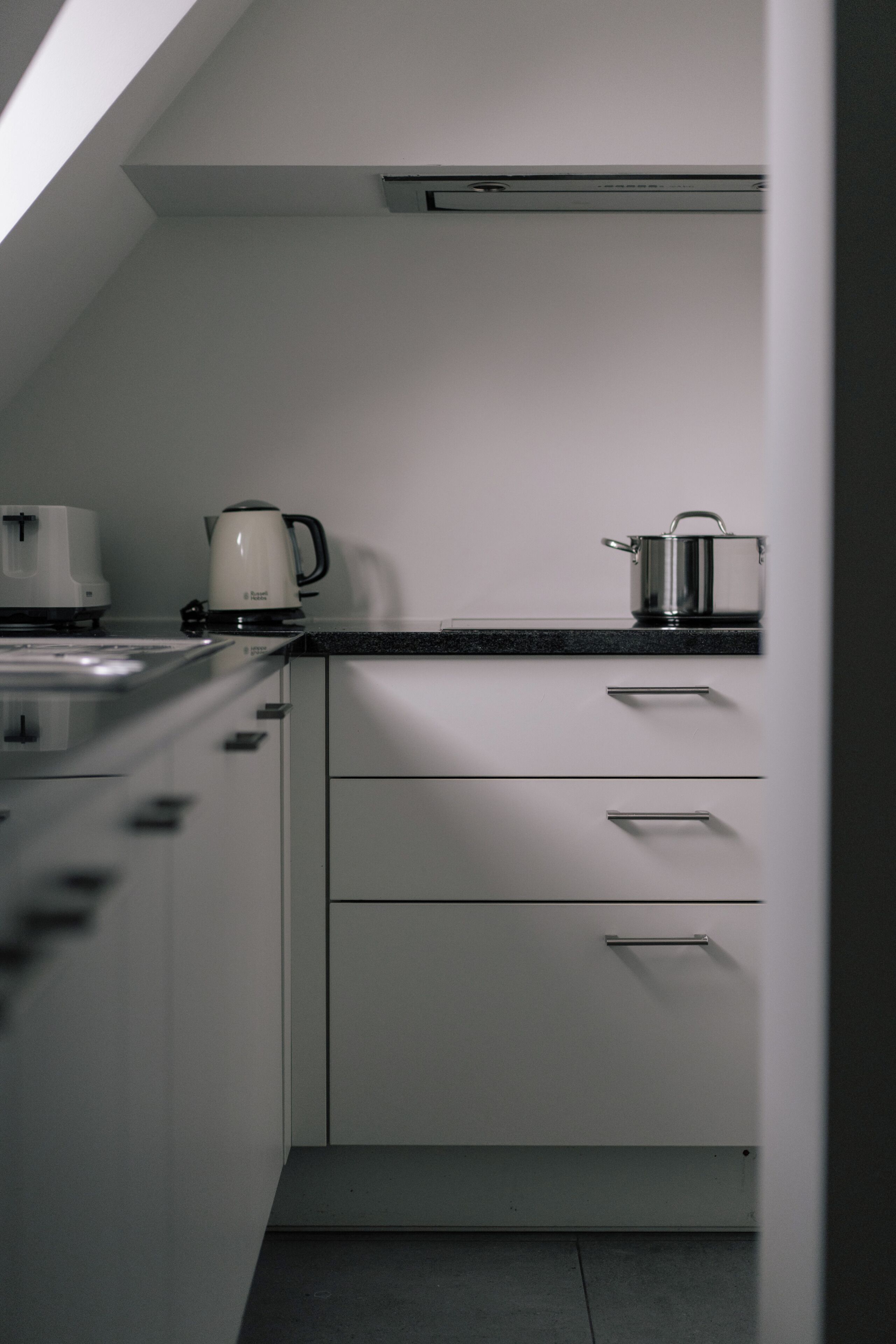 Una cocina con estufa, tetera y olla sobre la encimera, armarios blancos y un respiradero en el techo.